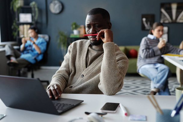 Портрет нудного афроамериканця, який грає з олівцем, працюючи за столом в офісі і дивлячись на екран ноутбука - Фото, зображення