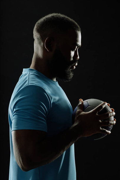Retrato dramático de vista lateral del jugador de fútbol afroamericano sosteniendo la pelota contra fondo negro con contorno - Foto, Imagen