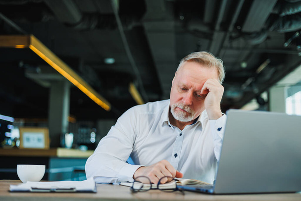 Ein grauhaariger Geschäftsmann sitzt an einem Laptop und blickt unglücklich auf das schwache Umsatzwachstum. Verluste im Unternehmen enttäuschen den Manager. - Foto, Bild