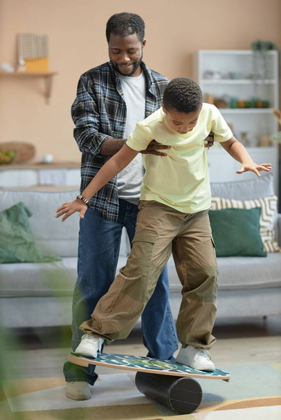 Vertikales Ganzkörperporträt von schwarzem jungen Vater, der seinem Sohn beibringt, auf dem Balance Board zu stehen und zusammen Spaß zu haben - Foto, Bild