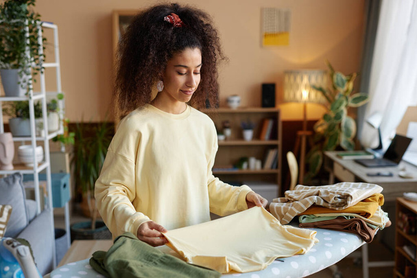 Retrato de cintura hacia arriba de una joven sonriente con gran pelo rizado doblando la ropa en la tabla de planchar en el espacio de copia en casa - Foto, imagen