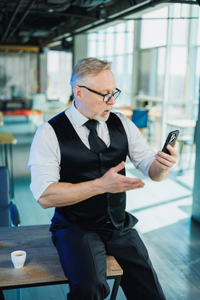 Un hombre de negocios barbudo de pelo gris sostiene un teléfono en sus manos, un hombre está viendo un video en el teléfono. Trabajo en la oficina - Foto, imagen