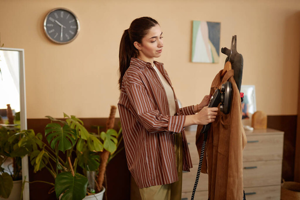 Widok z boku portret młodej kobiety przy użyciu parowca odzieżowego na aksamitnej kurtce w przytulnej przestrzeni kopia domu - Zdjęcie, obraz