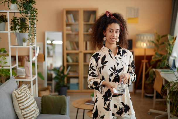 Portrait taille haute de jeune femme afro-américaine avec de gros cheveux bouclés souriant à la caméra à la maison et tenant minuscule arrosage peut copier l'espace - Photo, image