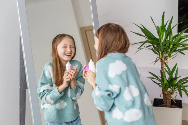 Happy preteen child applies facial cream while smiling at her reflection, with vibrant houseplant adding touch of greenery to scene. - Photo, Image