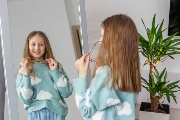 Nastoletnia dziewczyna eksperymentuje z przymierzaniem pary okrągłych okularów, jej uśmiech odbija się w lustrze sypialni z rośliną doniczkową w rogu. - Zdjęcie, obraz
