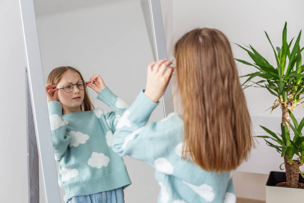 Ajattelevainen preteen tyttö säätää huolellisesti pari tyylikäs pyöreä lasit, tarkkailun hänen heijastus täyspitkä peili kotona. - Valokuva, kuva