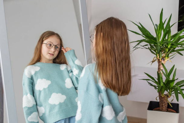 Zufrieden lächelnd probiert das Mädchen eine schicke runde Brille vor dem Spiegel an, deren dekorative Pflanze die heimelige Atmosphäre unterstreicht.. - Foto, Bild