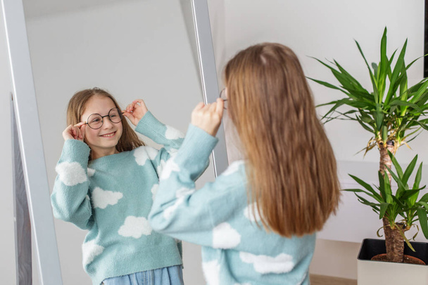 Sonriendo contenta, la adolescente se prueba un par de gafas redondas elegantes frente al espejo, con plantas decorativas que mejoran la atmósfera hogareña. - Foto, imagen