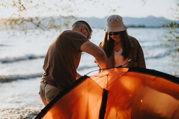 Młody mężczyzna i kobieta pracują razem nad rozbiciem pomarańczowego namiotu nad jeziorem, uchwycając radosny moment koleżeństwa i relaksu na świeżym powietrzu. - Zdjęcie, obraz