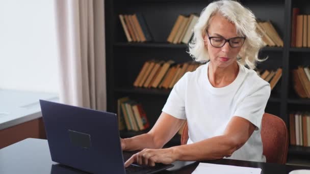 Αυτοπεποίθηση κομψή μεσήλικη ηλικιωμένη γυναίκα που χρησιμοποιεί φορητό υπολογιστή στο χώρο εργασίας. Μεγαλύτερη ώριμη γυναίκα επιχειρηματίας κάθεται στο τραπέζι του γραφείου με φορητό υπολογιστή και χαρτί έγγραφο. Αρχηγός εκπαιδευτικός επαγγελματίας εργαζόμενος - Πλάνα, βίντεο