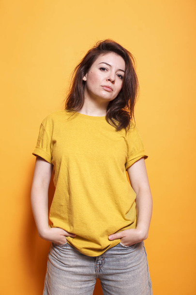 Confident woman with hands in pocket, brown hair, and casual clothing stands against an isolated orange background. Her expressive eyes and friendly demeanor are captivating. - Photo, Image