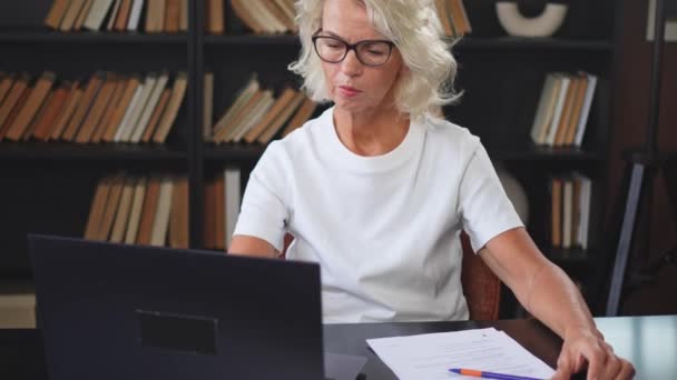 Starší žena středního věku, která na pracovišti píše poznámky na notebooku. Soustředěné dospělé staré podnikatelky pracují v kanceláři a vytvářejí písemné záznamy, které dělají online práci. Starší šéf profesionální pracovník - Záběry, video