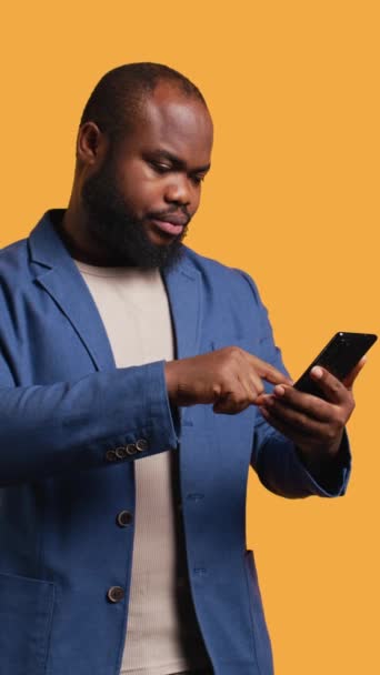 Vidéo verticale Portrait d'un homme joyeux tenant un téléphone portable célébrant, jetant des poings dans l'air et criant. Délicieux afro-américain lisant de bonnes nouvelles sur téléphone portable, acclamations, studio - Séquence, vidéo