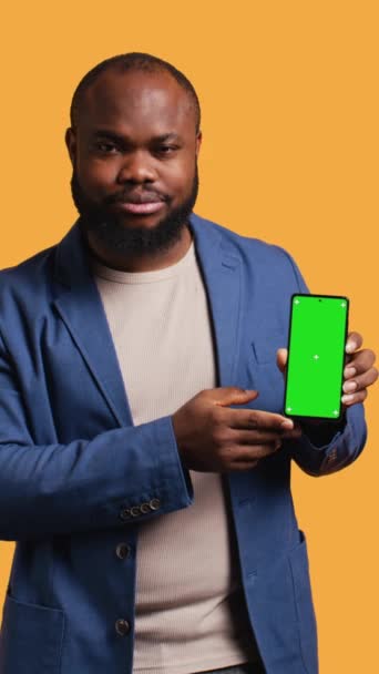 Вертикальное видео Расстроенный африканский американец показывает большой палец вниз знак жеста держа зеленый экран телефона, не одобряя. BIPOC человек делает отрицательный жест рукой в отношении смартфона, студийный фон - Кадры, видео