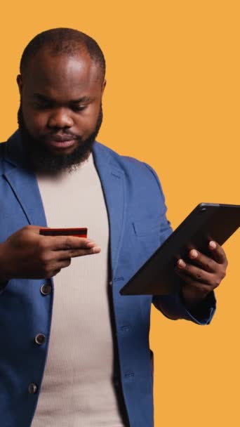 Vertical video Smiling man scrolling on tablet touchscreen, using internet and credit card to pay for products. African american person happily browsing online shopping websites, studio background - Footage, Video