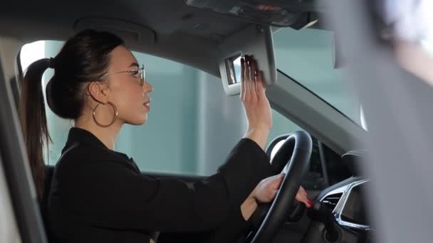 Een jonge vrouw in een auto is gevaarlijk aan het multitasken, kijkt naar haar telefoon, brengt lippenstift aan, praat aan de telefoon, controleert de achteruitkijkspiegel, creëert een chaotische en riskante situatie - Video