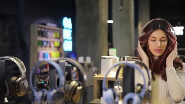 Молода дівчина насолоджується музикою в нових навушниках в цифровому магазині, дивлячись на камеру. Купівля навушників для прослуховування музики, для роботи. - Кадри, відео