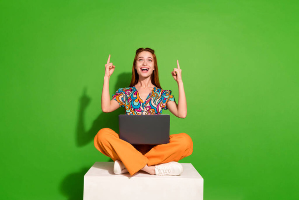 Ganzkörper-Porträt von hübschen jungen Mädchen sitzen Podium Laptop direkte Finger nach oben leeren Raum tragen Top isoliert auf hellgrünem Hintergrund. - Foto, Bild
