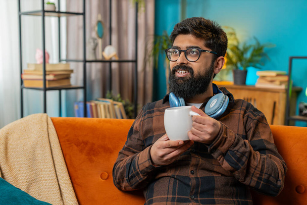 Hymyilevä iloinen intialainen nuori mies juo kupin lämmintä kahvia tai yrttiteetä istuu kotona olohuoneen sohvalla aamulla. Latinomies nauttii rentoutumisesta yksin. Rentoutuminen, tauko. - Valokuva, kuva
