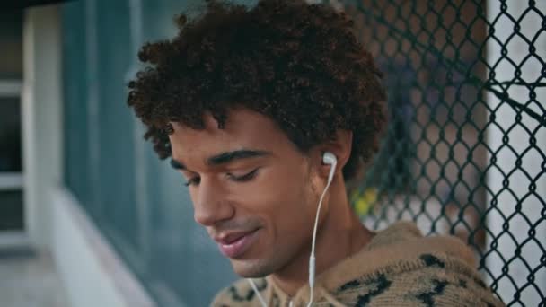 Smiling teenager listening song in headphones at quiet urban area closeup. Relaxed latino zoomer enjoy favorite music in earphones standing street fence. Stylish curly guy relaxing at leisure time. - Footage, Video