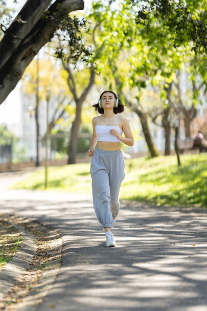 Μια γυναίκα τρέχει σε ένα μονοπάτι σε ένα πάρκο. Φοράει ακουστικά και ακούει μουσική. - Φωτογραφία, εικόνα