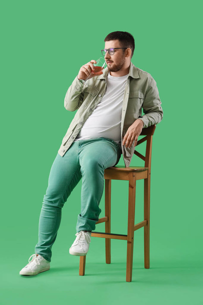 Νεαρό σομελιέ δοκιμάζοντας κρασί ενώ κάθεται σε ξύλινη καρέκλα με φόντο το πράσινο - Φωτογραφία, εικόνα