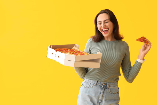 Happy young woman holding cardboard box with tasty pizza on yellow background - Photo, Image