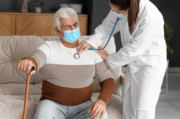 Junge Krankenschwester in medizinischer Maske hört Senior zu Hause mit Stethoskop - Foto, Bild