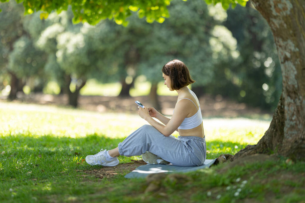 Una mujer está sentada debajo de un árbol y mirando su teléfono. Ella lleva una camiseta blanca y pantalones grises - Foto, imagen