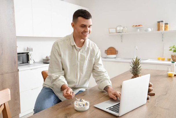 Junger Mann mit leckerem Joghurt mit Laptop in Küche - Foto, Bild