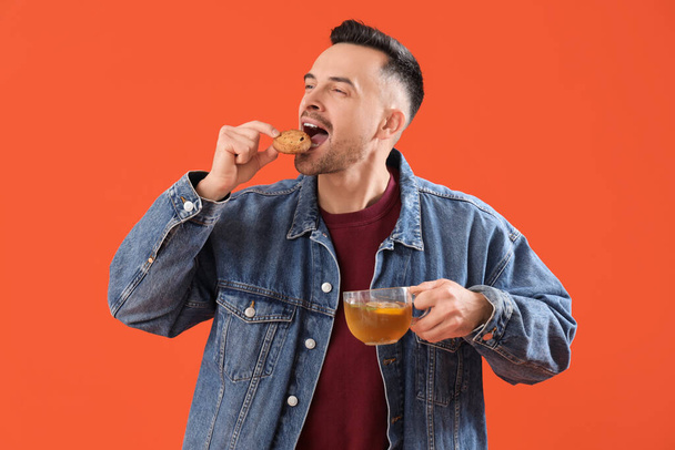 Knappe man met een kop citroen thee etend koekje op oranje achtergrond - Foto, afbeelding
