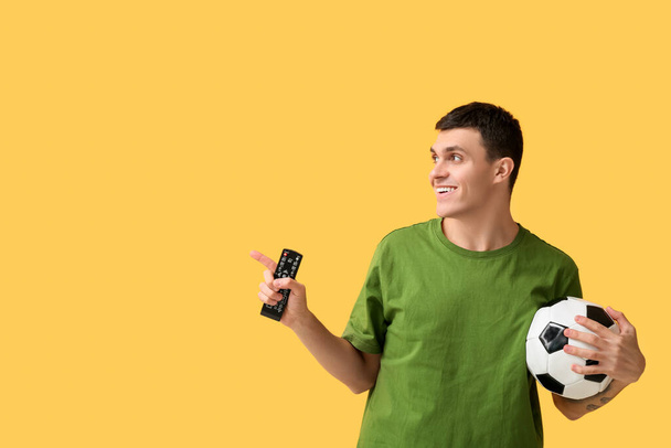 Jeune homme avec ballon de football et télécommande TV pointant quelque chose sur fond jaune - Photo, image