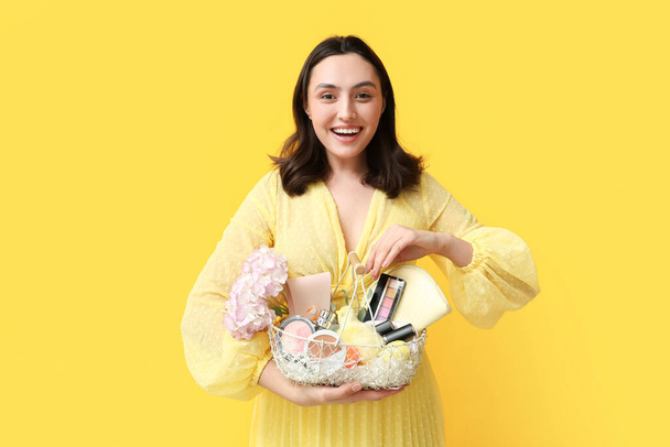 Happy young woman holding basket with decorative cosmetics and Easter decor on yellow background - Photo, Image