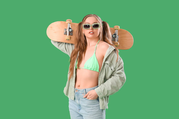 Красива молода жінка в стильному вбранні зі скейтбордом на зеленому фоні - Фото, зображення