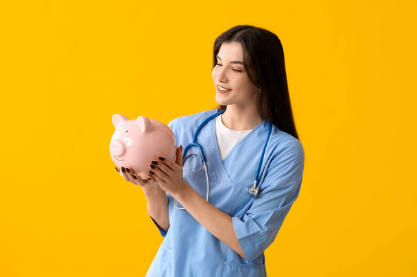 Mooie jonge vrolijke vrouwelijke arts met spaarvarken bank en stethoscoop op gele achtergrond. Gezondheidszorgconcept - Foto, afbeelding