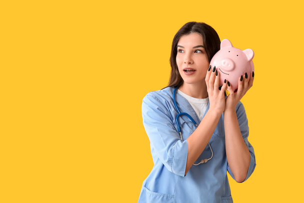 Beautiful young shocked female doctor with piggy bank and stethoscope on yellow background. Health care concept - Photo, Image