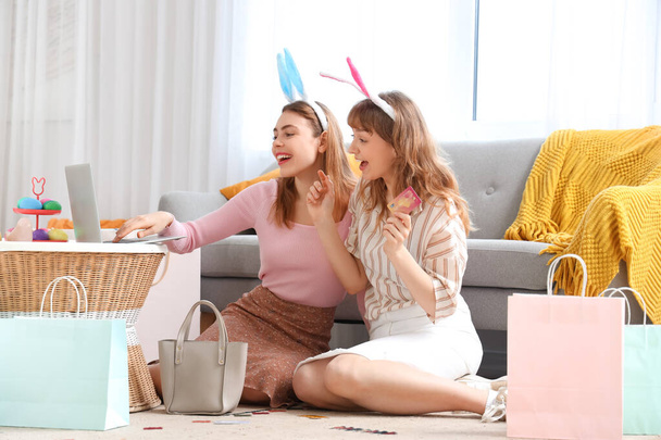 Счастливые молодые женщины в головных уборах кроликов с ноутбуком, кредитными карточками и бумажными пакетами на Пасху сидят дома на полу. Интернет-магазины - Фото, изображение