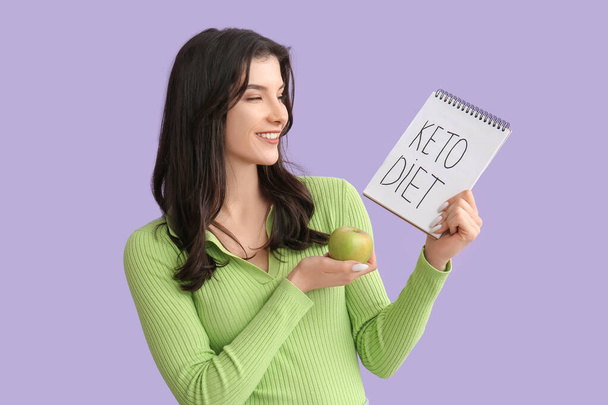 Feliz joven sonriente sosteniendo un cuaderno con texto KETO DIET y manzana sobre fondo lila - Foto, imagen