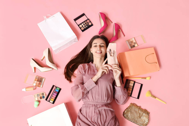 Joyeux sourire jeune femme avec des pinceaux de maquillage, des chaussures et des palettes de fards à paupières couché sur fond rose - Photo, image