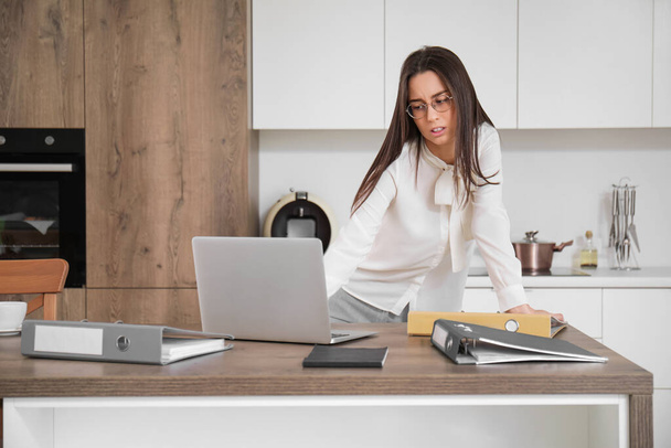 Jeune femme d'affaires fatiguée avec ordinateur portable et dossiers de bureau sur la table dans la cuisine - Photo, image