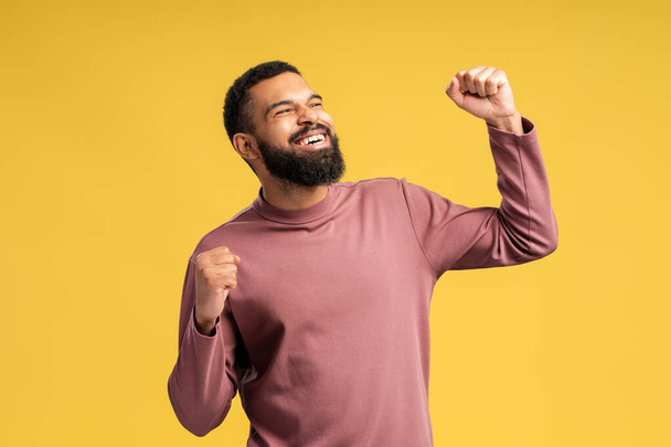 Usmívající se Afroameričan s plnovousem dělá vítězný gesto, pózuje ve studiu, izolován na žlutém pozadí. Lidé emoce životní styl koncept - Fotografie, Obrázek