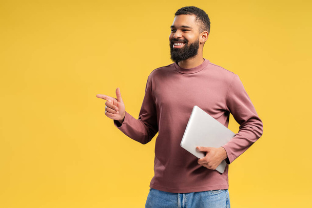 Happy African American bearded man looking to the side in studio, holding laptop and pointing to copy space by finger, isolated on yellow background. Online technology concept - Photo, Image