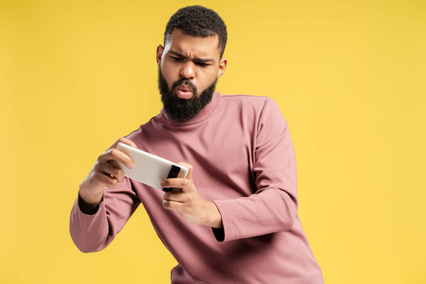 Uomo afroamericano in abiti casual mentre fa foto, utilizzando smartphone, guardando lo schermo, isolato su sfondo giallo. Concetto di lifestyle - Foto, immagini