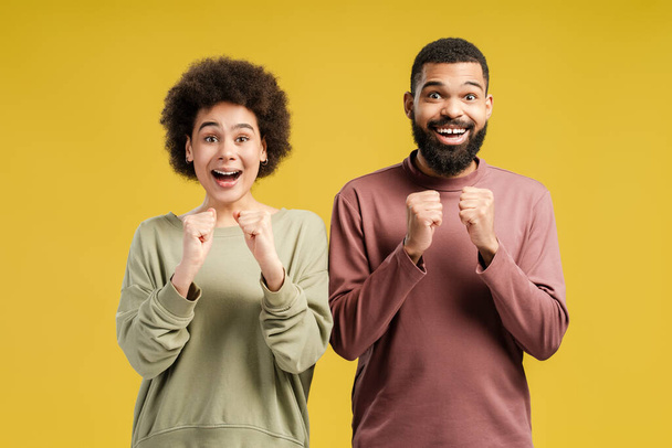 Smiling African American man and woman looking at camera while having triumph, celebrating, isolated on yellow background. Lifestyle people concept - Photo, Image