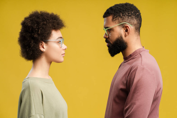 Giovane uomo e donna afroamericani che si guardano in studio, isolati su sfondo giallo. Vista laterale. Stile di vita concetto di persone - Foto, immagini