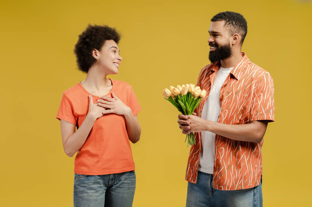 Schöner afroamerikanischer Mann überreicht seiner geliebten Frau Rosen, isoliert auf gelbem Hintergrund. Urlaub, Gruß und Menschen-Konzept - Foto, Bild