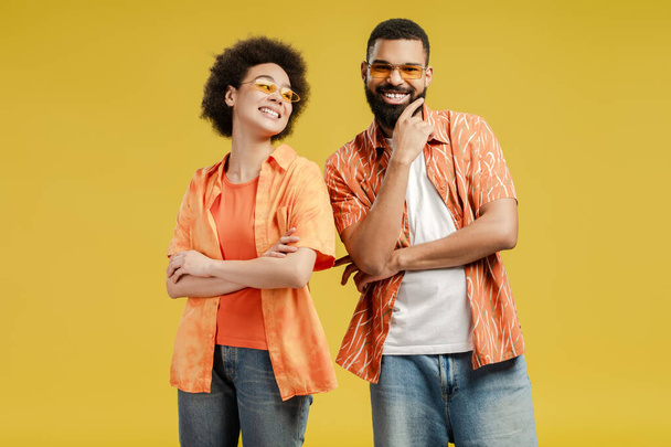 Smiling African American woman looking at man, wearing eyeglasses, posing in studio, isolated on yellow background. Vacation, summer party concept - Photo, Image