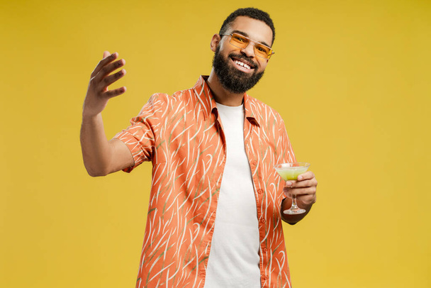 Hombre afroamericano sonriente en gafas con cóctel de verano, acogedor, posando en el estudio, mirando a la cámara, aislado en amarillo. Vacaciones, concepto de fiesta de verano - Foto, Imagen