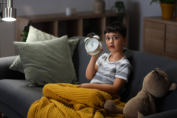 Lindo niño pequeño con oso de peluche y reloj despertador sentado en el sofá en la sala de estar - Foto, Imagen